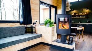 A modern living room with a built-in wood stove, a grey sofa, wooden flooring, and a view of the outdoors through a large window. - Nature lodge Sweltsje, Akkrum - sleeps 4 by the water (Akkrum)