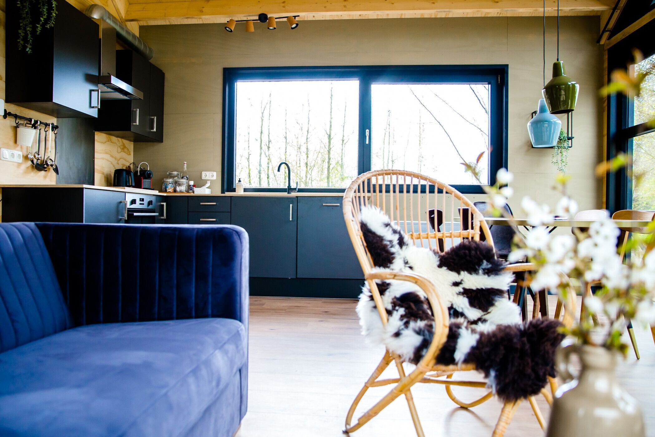 A modern kitchen with dark cabinetry, a large window, and a dining area with a blue sofa and a wicker chair.