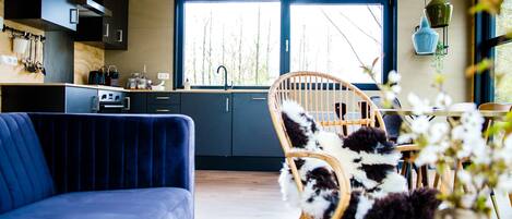 A modern kitchen with dark cabinetry, a large window, and a dining area with a blue sofa and a wicker chair.