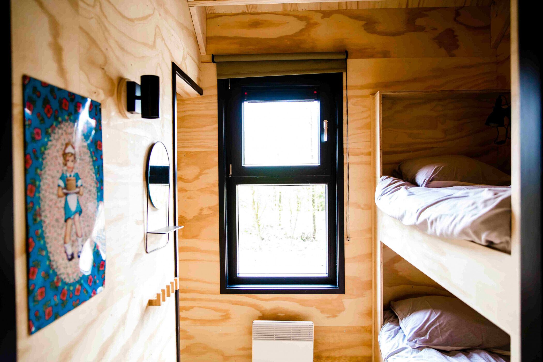 Een kleine, compacte kamer met een stapelbed, een raam met uitzicht op bomen, een wandairconditioner en een lijst met een foto.