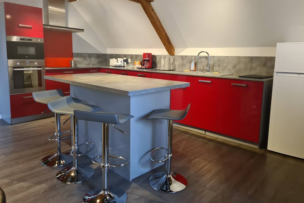 A modern kitchen with red cabinets, a central island with bar stools, stainless steel appliances, and a wooden ceiling.