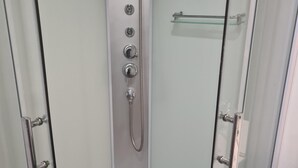 Une cabine de douche fermée par des parois en verre, dotée d’un pommeau de douche à main, d’un panneau de commande fixé au mur et d’une étagère en verre.