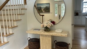 Un couloir avec un escalier en bois, un miroir rond, une console sur laquelle est posé un vase de fleurs et des paniers en osier.
