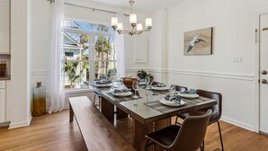 Un coin repas avec une table en verre, un banc en bois et des chaises. Un lustre est suspendu au-dessus de la table, et une fenêtre avec des rideaux se trouve sur le côté gauche.