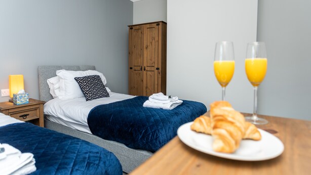 A hotel room with two beds, a wooden wardrobe, a bedside table with a lamp, and a breakfast setup with croissants and orange juice. - HC Property - The Old Clubhouse (Bishop Auckland)