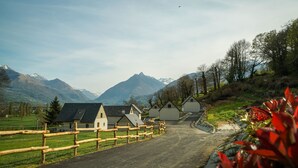 A rural road leading to a row of white buildings with sloped roofs, surrounded by mountains and a wooden fence. - Chalet Viscos (Les Chalets du Balandrau) (Ayzac-Ost)