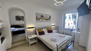 A modern bedroom with a large bed, a smaller bed in an adjacent room, a window with a view, and a unique ceiling light fixture. - Family Room inside the Guesthouse on the Arch (Monterosso al Mare)