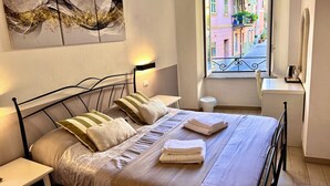 A neatly made bed with pillows and folded towels, a bedside table, a wall-mounted artwork, a window with a view of colorful buildings, and a small round table with a lamp. - Family Room inside the Guesthouse on the Arch (Monterosso al Mare)