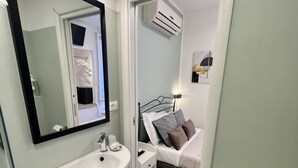 A modern bathroom with a white sink, mirror, and toilet. There is a bed with a white and brown bedspread in the adjacent room. - Camera Singola All'interno di un Affittacamere in Pieno Centro Storico (Monterosso al Mare)