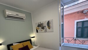 A bedroom with a bed, bedside table, air conditioning unit, artwork, and a window with a balcony. - Camera Singola All'interno di un Affittacamere in Pieno Centro Storico (Monterosso al Mare)