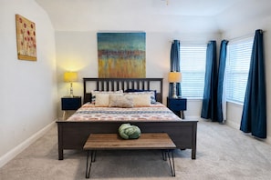 A bedroom with a bed, bedside tables, lamps, a bench, a large painting, and a window with curtains.