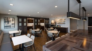 Een modern restaurant met houten vloeren, witte tafels en grijze stoelen. Er is een keuken met een bar en een zithoek met een grote houten tafel.