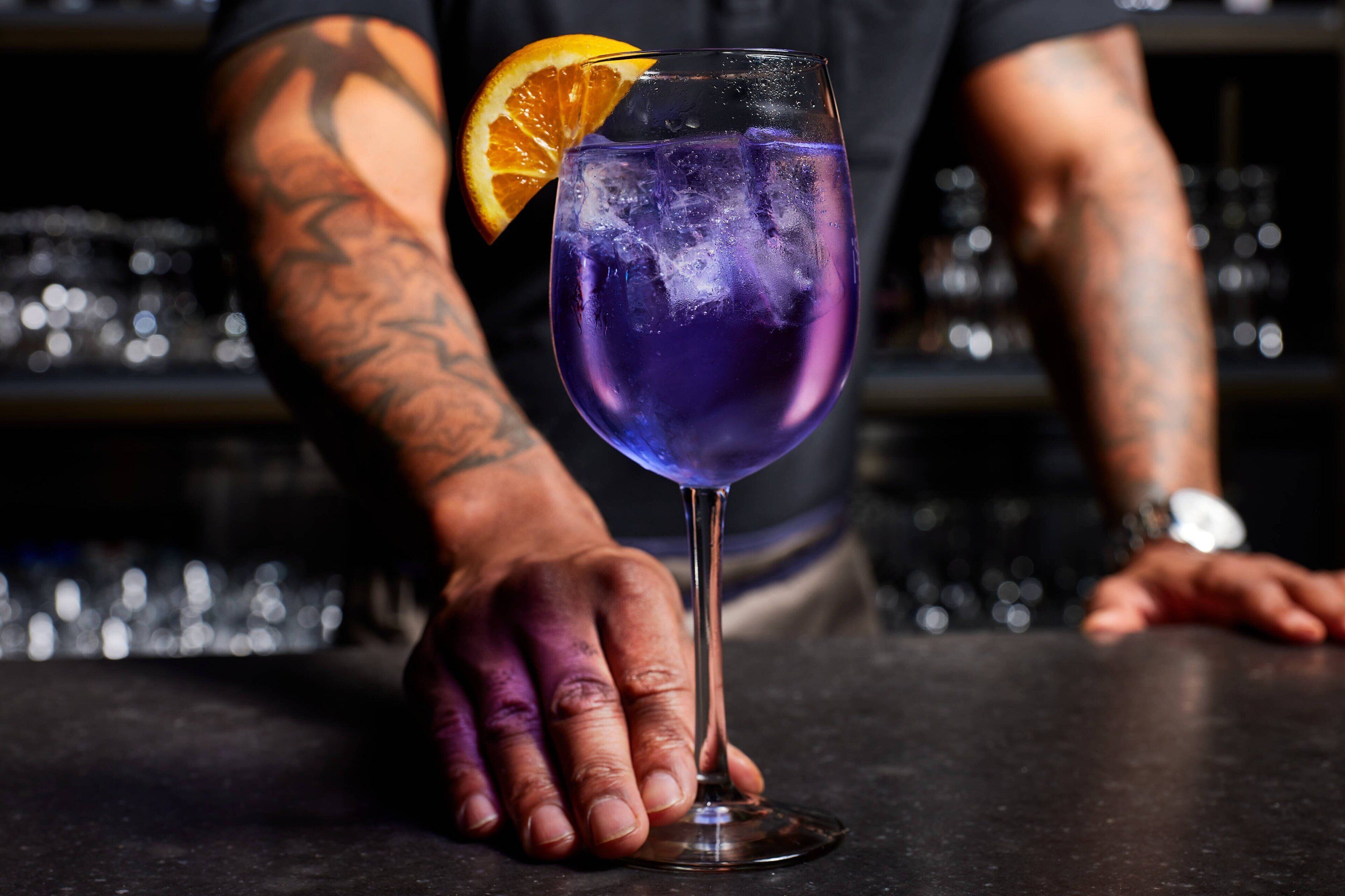 Une personne tenant un verre contenant une boisson violette, des glaçons et une tranche d’orange en guise de décoration.