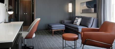 A modern hotel room with a desk, office chair, sofa, ottoman, and a standing chair.