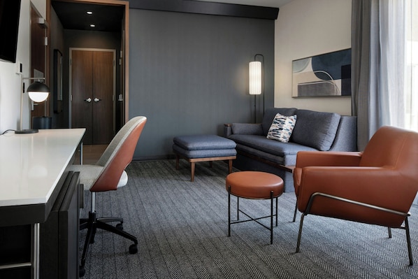 A modern hotel room with a desk, office chair, sofa, ottoman, and a standing chair. - Courtyard by Marriott Fresno Clovis (Clovis)