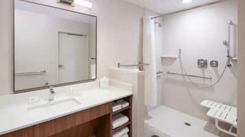 A modern bathroom with a large mirror, a sink, a shower with a grab bar, and a bench.