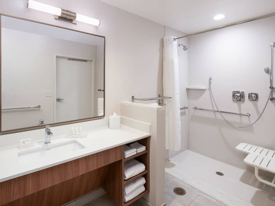 A modern bathroom with a large mirror, a sink, a shower with a grab bar, and a bench.