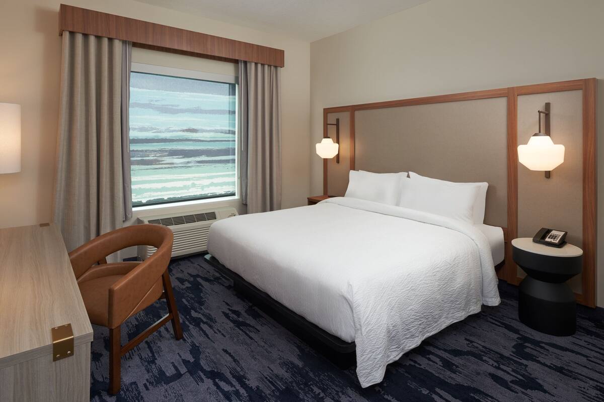 A hotel room with a large bed, a desk, a chair, a bedside table with a phone, and a window with a view of the ocean.