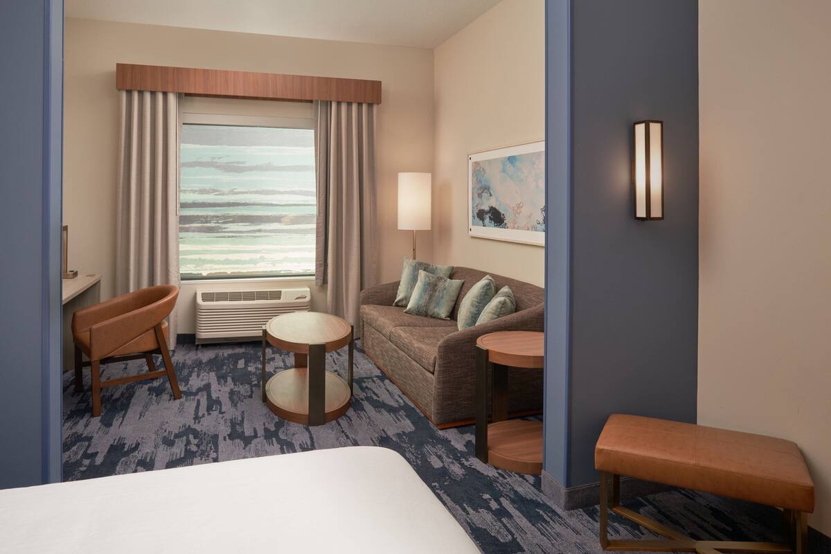 A hotel room with a large bed, a sofa, a small table, a bench, a desk, and a window with curtains.