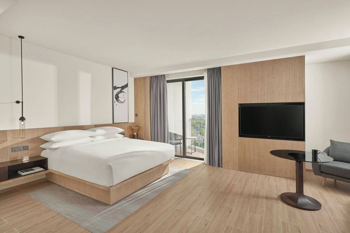 Quarto de hotel moderno com uma cama grande, uma TV, uma mesinha e uma varanda com cortinas.