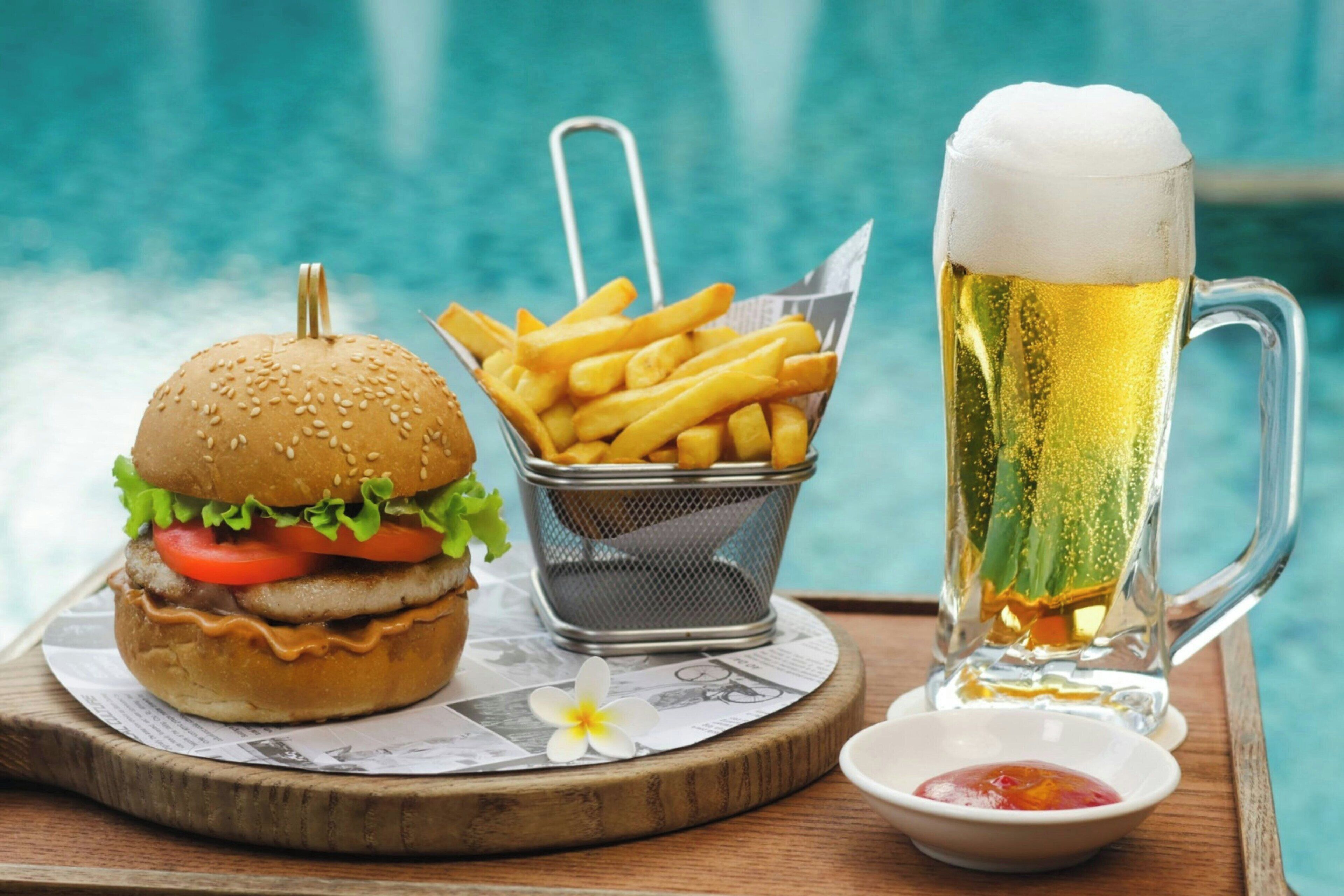 Un plateau repas avec un hamburger, des frites et une chope de bière, posé près d’une piscine.