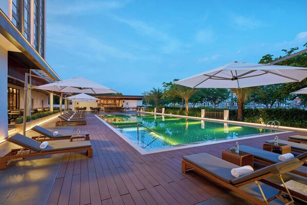 A rooftop pool area with sun loungers and umbrellas, surrounded by a wooden deck and a building with a bar/lounge area in the background.