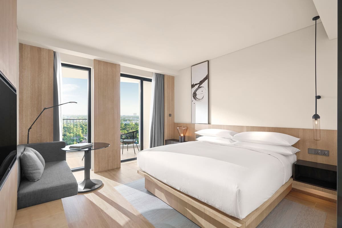 Quarto de hotel moderno com uma cama grande, um sofá, uma mesa pequena e uma varanda com vista para a cidade.