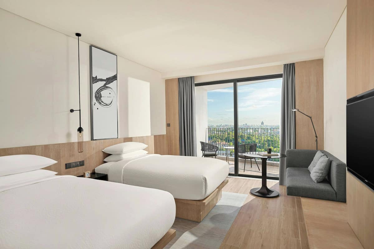 Quarto de hotel com duas camas, um sofá, uma TV e varanda com vista para a cidade.