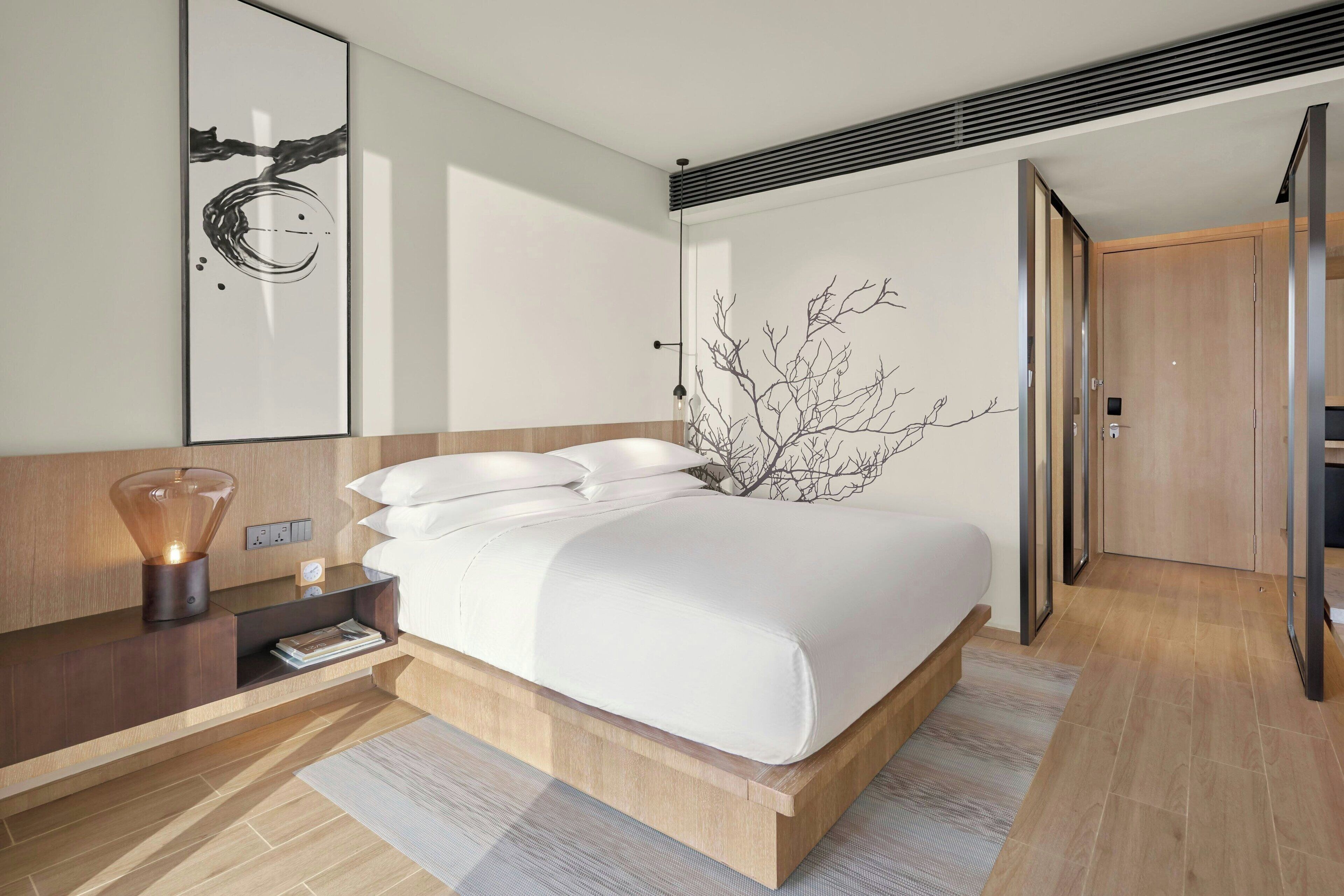 A modern bedroom with a large bed, a bedside table with a lamp, and a wall-mounted artwork.