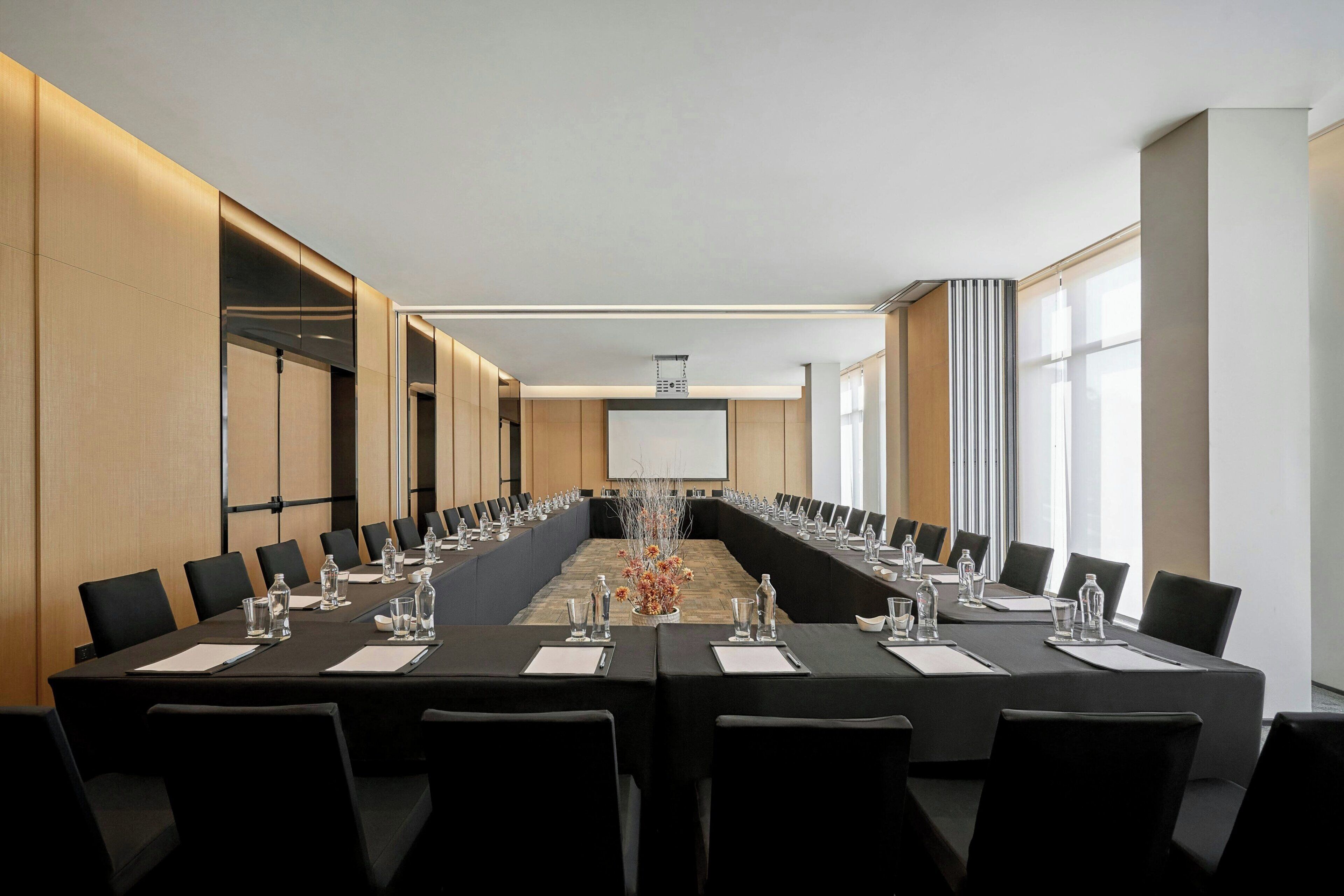 A conference room with a long table, black chairs, and a projector screen at the front.
