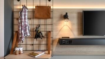Una sala de estar moderna con un televisor montado, una lámpara de techo, un estante de madera con un libro y dos latas, y un perchero con una chaqueta y un objeto decorativo.
