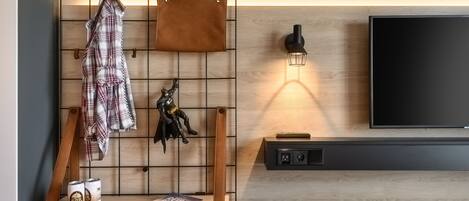A modern living room with a mounted TV, a wall-mounted light fixture, a wooden shelf with a book and two cans, and a wall-mounted rack holding a jacket and a decorative item.