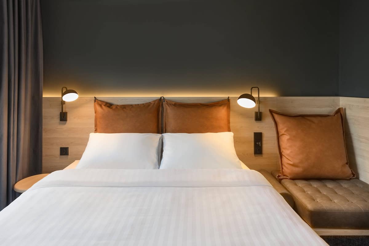 A hotel room with a large bed, two bedside lamps, a wooden headboard, and a small sofa with a pillow.
