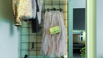 A room with a mirror reflecting a bed and a wall with a grid pattern. Clothes are hanging on a rack, and a pair of shoes is on the floor.
