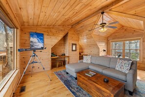 Interior de cabana de tronco com teto de madeira, um telescópio, um sofá cinzento e uma grande janela.
