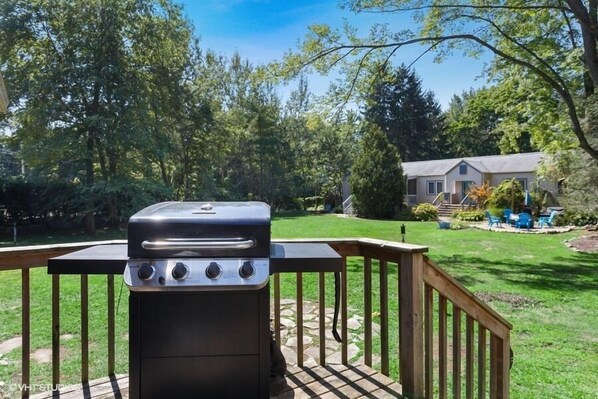 Un barbecue sur une terrasse en bois, avec un chemin de pierres menant à une maison située dans une zone boisée.