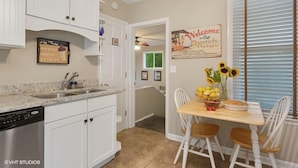 A kitchen with white cabinets, a granite countertop, a dishwasher, and a dining area with a wooden table and chairs. There is a sign on the wall that says "Welcome to Our Beach House." - Union Pier Home With Hot Tub and One Block To Beach! (Union Pier)