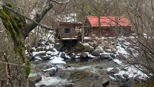 A rustic cabin with a red roof, surrounded by a forested area with a stream flowing through rocks and snow patches. - French Creek Inn.    Creekside Cabin with Fire Sauna (Riggins)