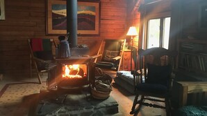 A cozy cabin interior with a wood-burning stove, a painting on the wall, and a window providing natural light. - French Creek Inn.    Creekside Cabin with Fire Sauna (Riggins)