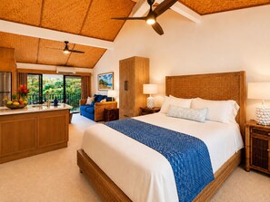 A hotel room with a large bed, a wooden headboard, a balcony with a view of greenery, a kitchenette, and a ceiling fan. - Princeville AC + Purple King Top-Floor Tiki Suite, Near 1 Hotel & Beaches (Princeville)