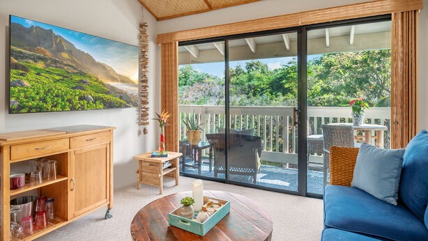 Un balcón amplio con vistas a zonas verdes y montañas, amueblado con un sofá azul, una mesa de madera y una silla de mimbre.