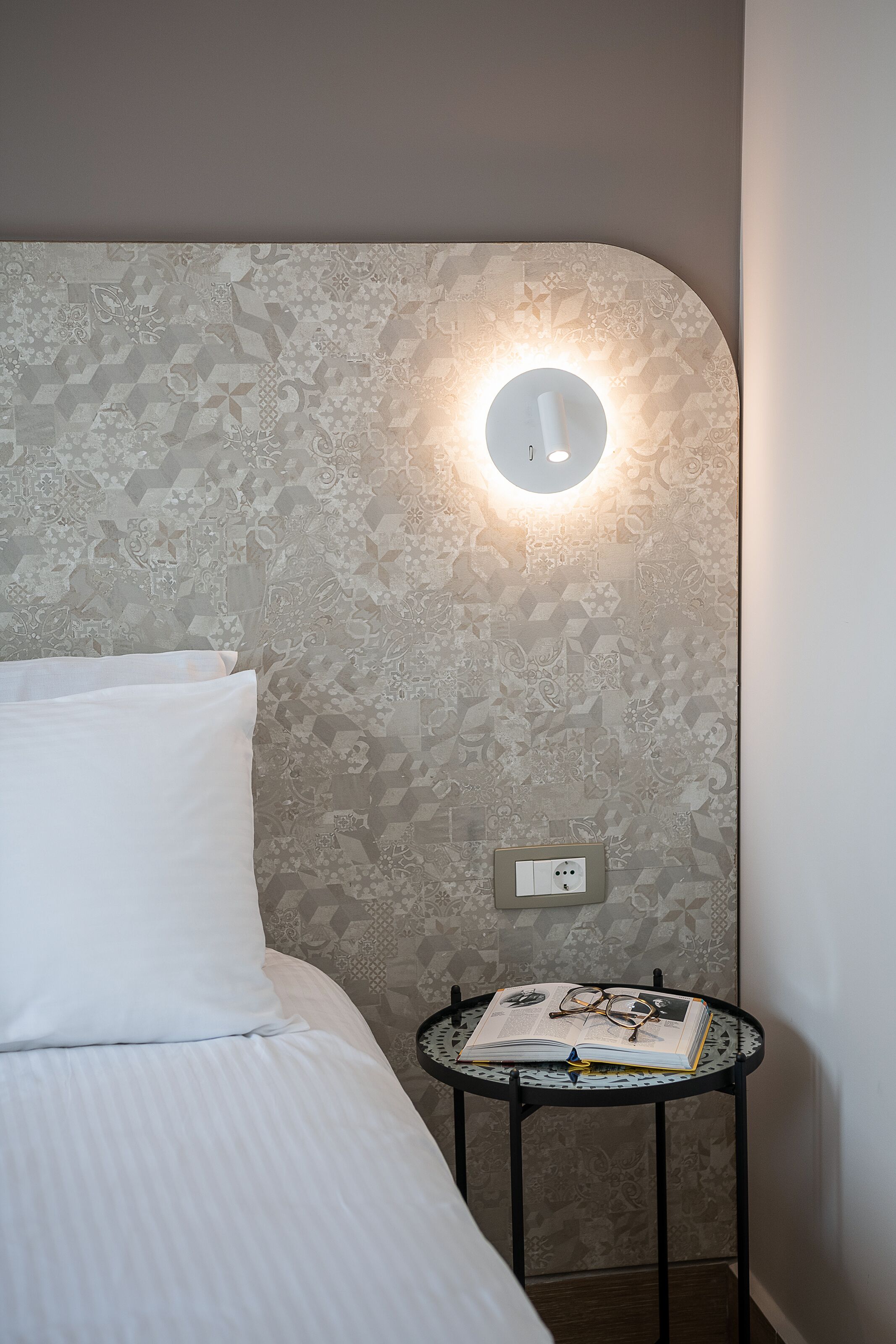Un lit bien fait, avec un oreiller blanc et une table ronde à côté, sur laquelle on peut voir un livre et des lunettes.