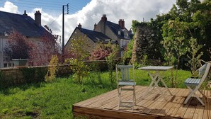 Una terraza de madera con una mesa y dos sillas, rodeada de vegetación exuberante y un muro de piedra.