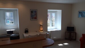 Una habitación con una mesa de comedor, sillas, un hervidor, una lámpara, un televisor y dos ventanas.