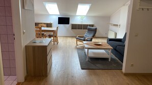 Un salon moderne comprenant un coin repas, un canapé gris, une table basse en bois et un téléviseur fixé au mur.