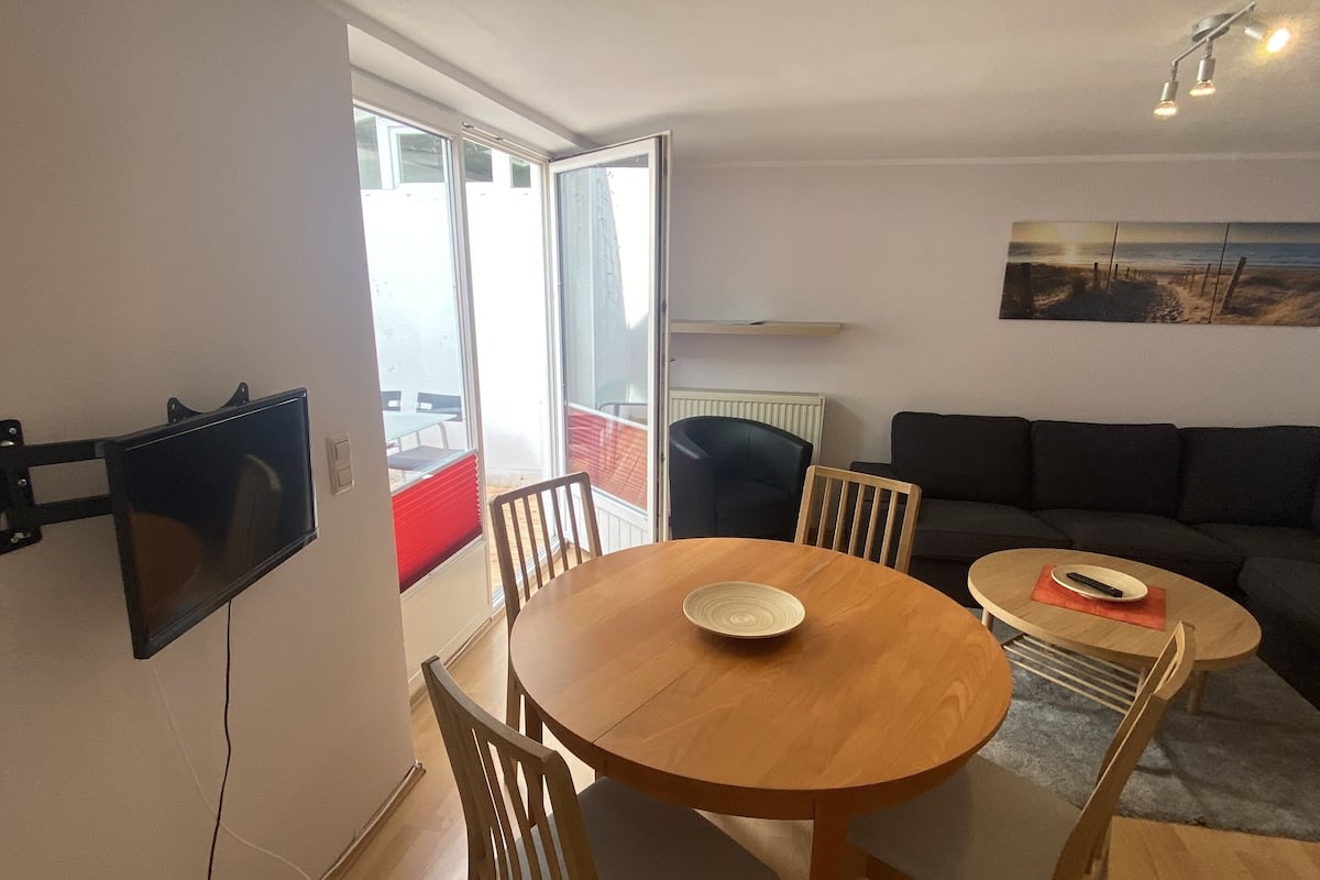 Un comedor con una mesa redonda de madera, cuatro sillas, un televisor de pantalla plana en la pared y un cuadro sobre el sofá.