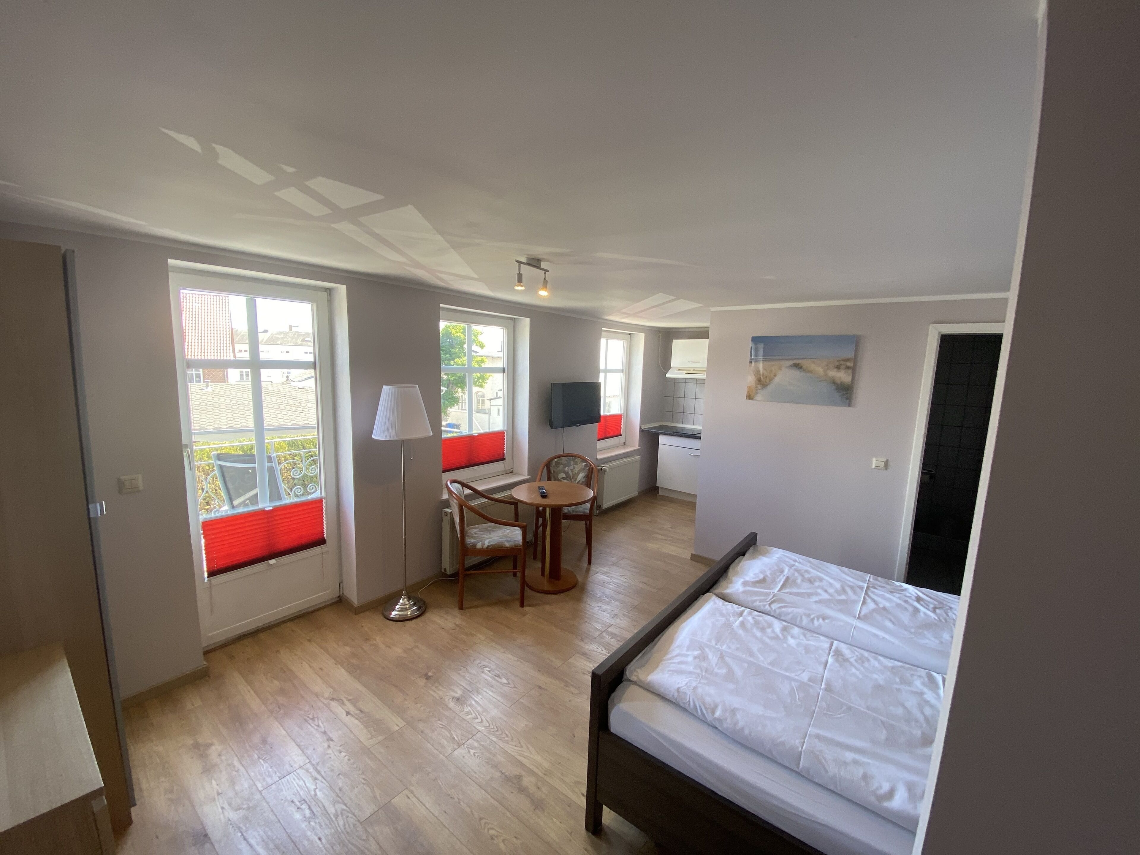 A bedroom with a bed, a small dining area with a table and chairs, a window with red blinds, and a kitchenette.