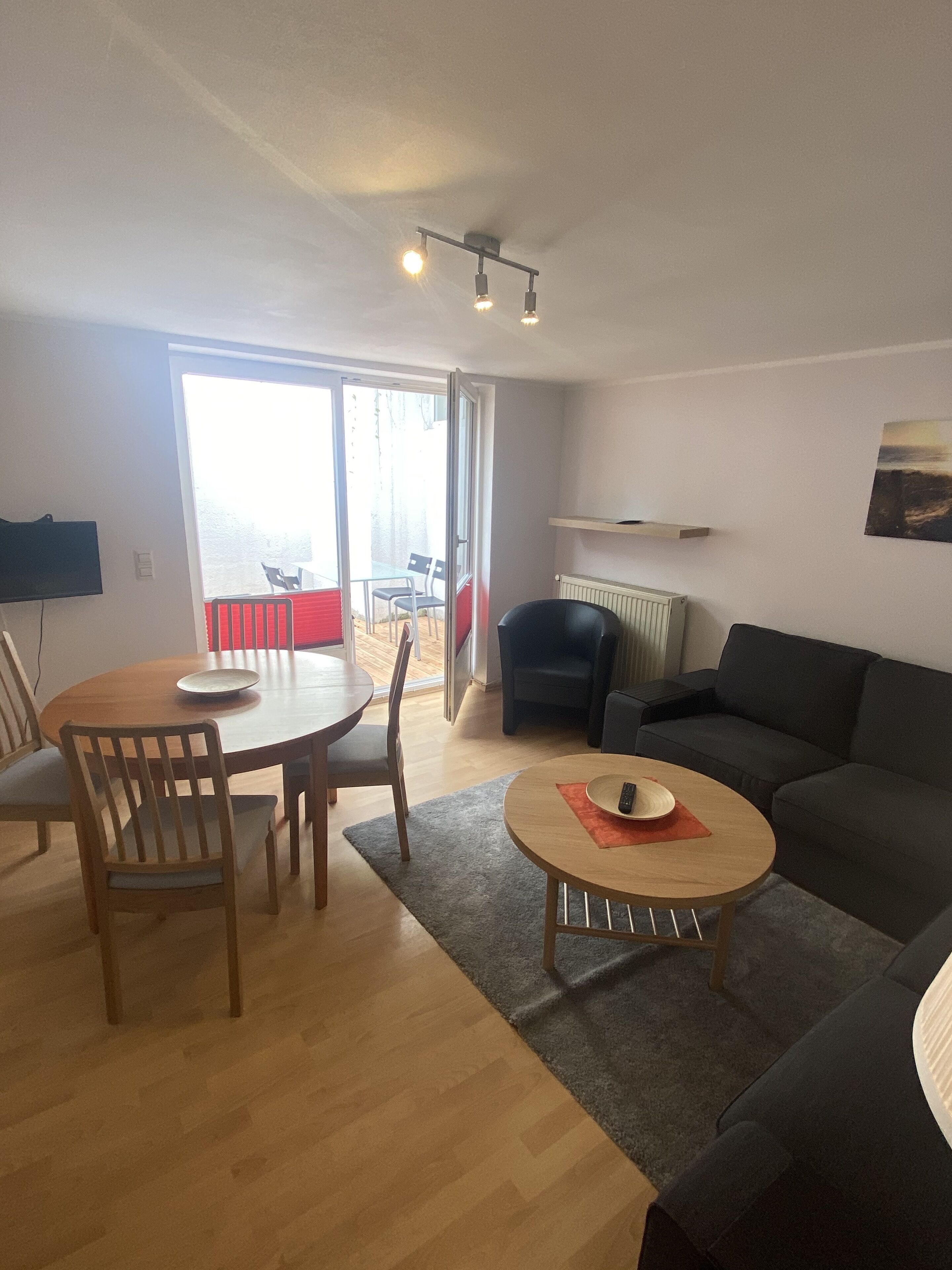 A living room with a dining area, a sofa, a TV mounted on the wall, and a large window.