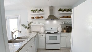 Una cocina con gabinetes blancos, una estufa blanca y un lavamanos. Hay plantas en macetas en los estantes y una alfombra en el suelo.