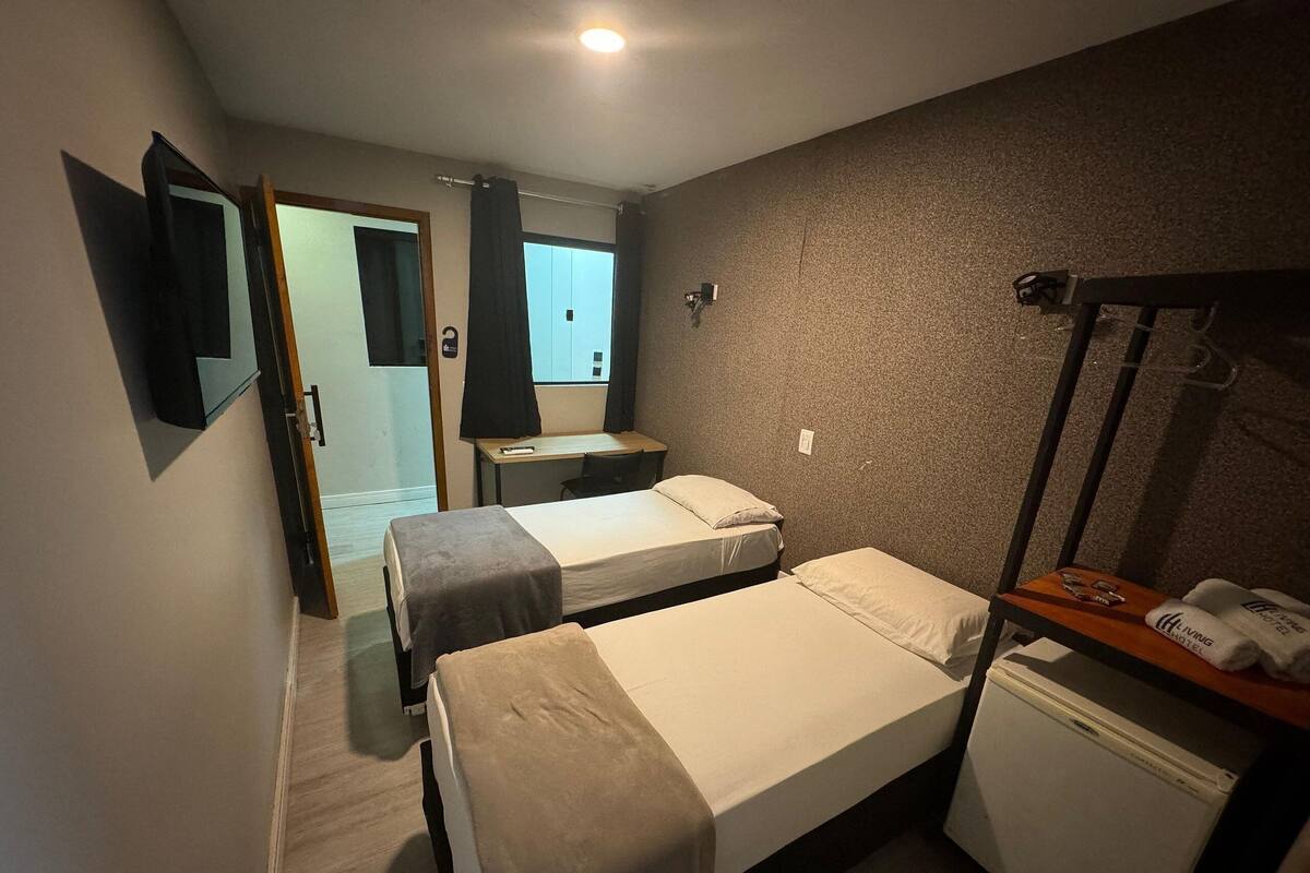 A small, dimly lit hotel room with two single beds, a desk, a mirror, and a small refrigerator.